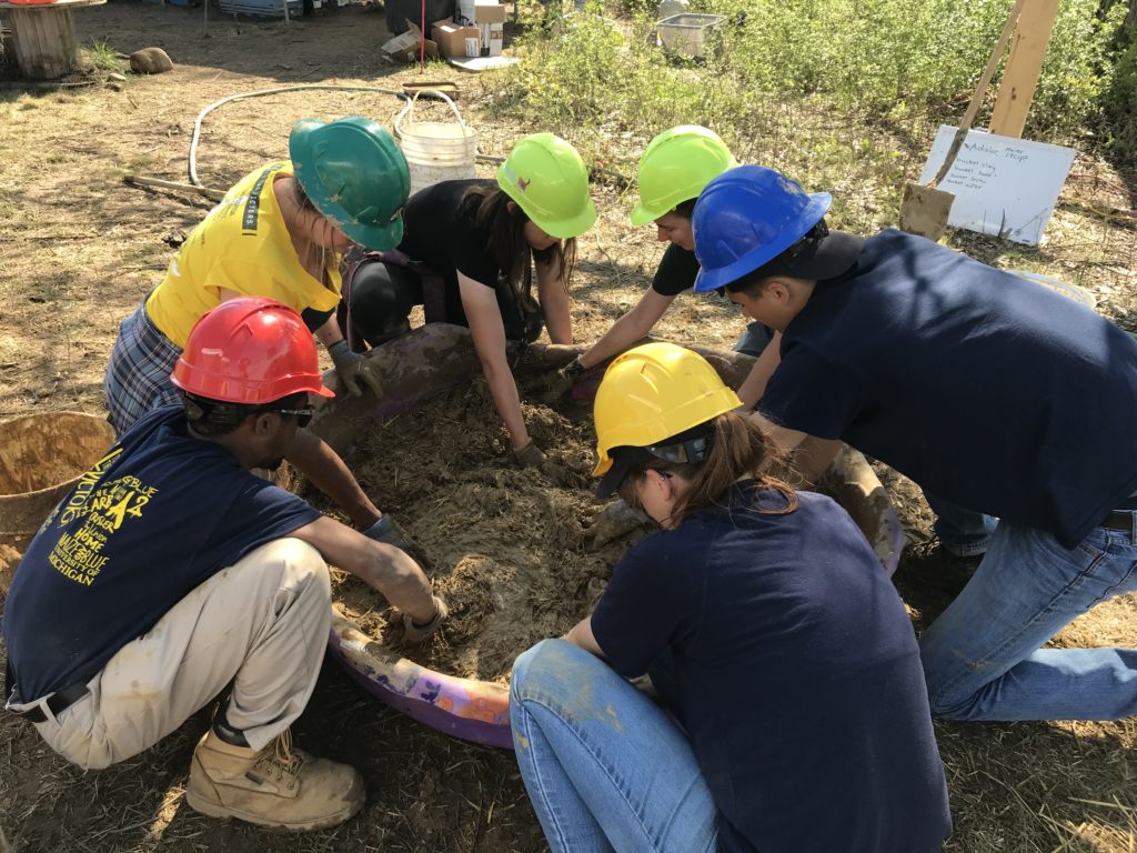 Six students mixing adobe, wearing multicolored safety helmets.