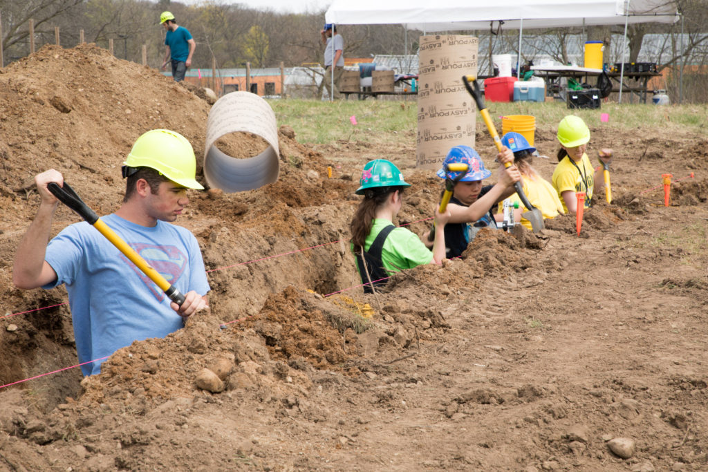 Five students in multicolored construction helmets stand in man-made ditch digging the foundation for the building