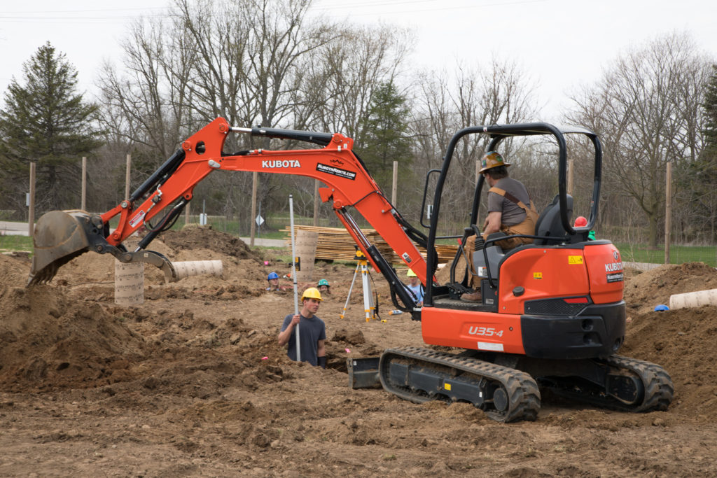 Man sitting in excavator converses with another man digging in a hole that has been dug nearby