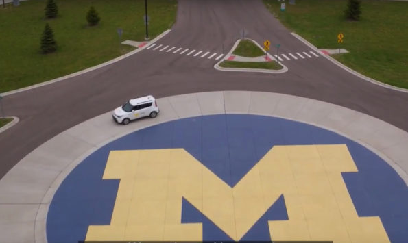 An aerial shot of a white vehicle driving along a sidewalk that encases a large block M.