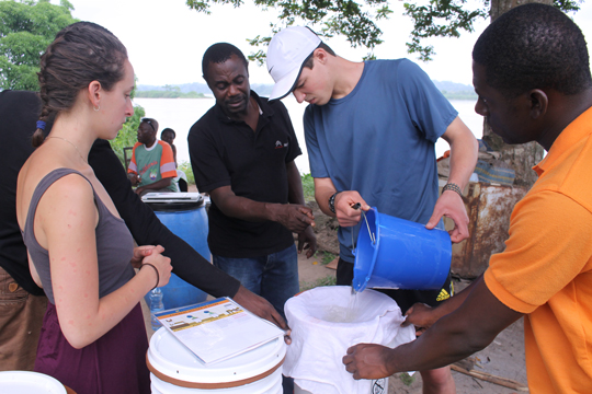 A student pours water into a water filter as a few people stand around watching him