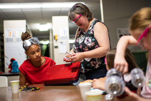 Facilitator demonstrates the use of a tool for two students in the background, while another student experiments with a toy in the foreground. All are wearing safety goggles.