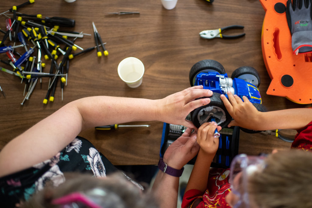 A facilitator and student are seen from above, both working on a toy car with large wheels, tools scattered on the table in front of them.