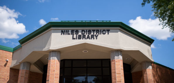 The outside of the Niles District Library, two brick columns, a green roof, and a large sign displaying the library's name.