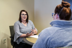 A woman (shown from front) sits across from another woman (shown from back) while they perform a mock interview.