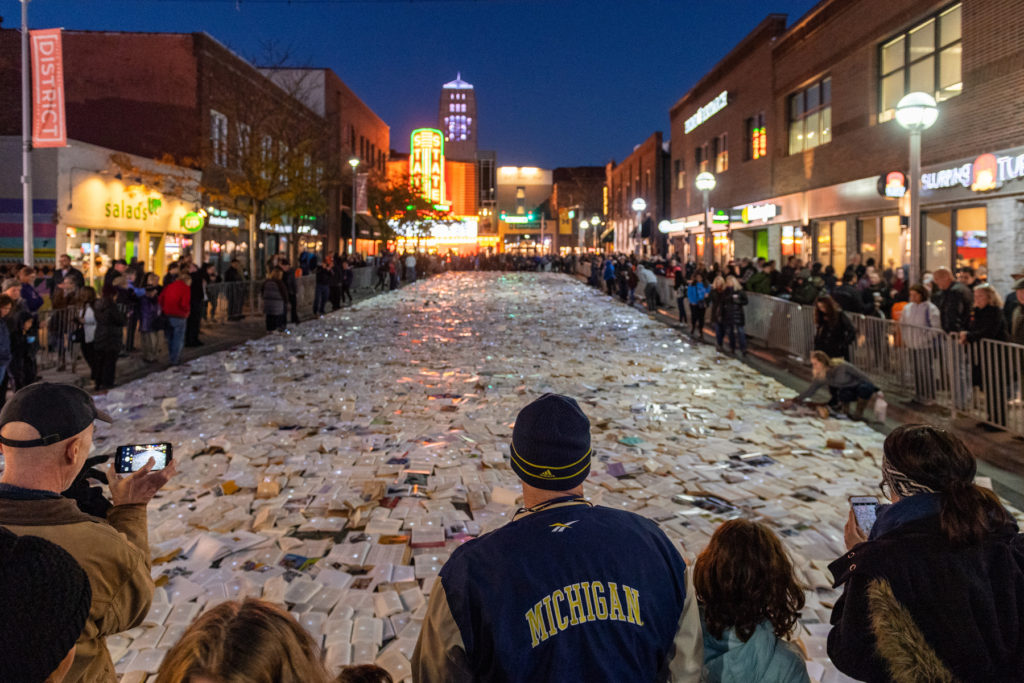 Large crowd stands around illuminated books on Liberty street at night, some taking pictures with their phones. State theater is pictured in the distance.