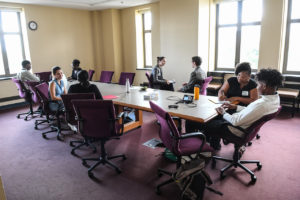 Many people sit around a large conference table in a room lit by natural light. The people are separated into pairs and are discussing amongst themselves.