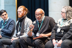 Wilkins speaks into a microphone while other speakers sit around him.