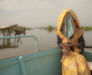 A woman in yellow with her face covered by cloth sits outside, a body of water in the background.