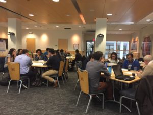 Students, faculty and staff are seated at various tables around a large room.