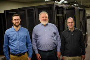 Three men stand side by side smiling in front of machinery.