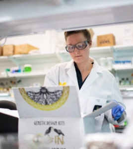 Nye wears safety goggles and a white lab coat while looking in a box of genetic design kits.