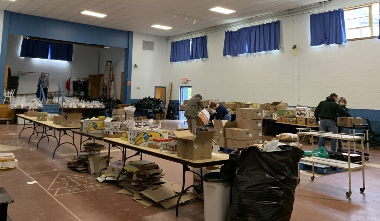 (A small gymnasium filled with opened and unopened cardboard boxes sitting on tables.)