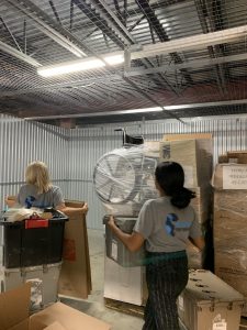 Two people carry boxes through a storage area with many stacked boxes.