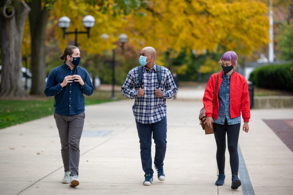 Three people walk side by side outside at the U-M campus wearing face masks and talking.