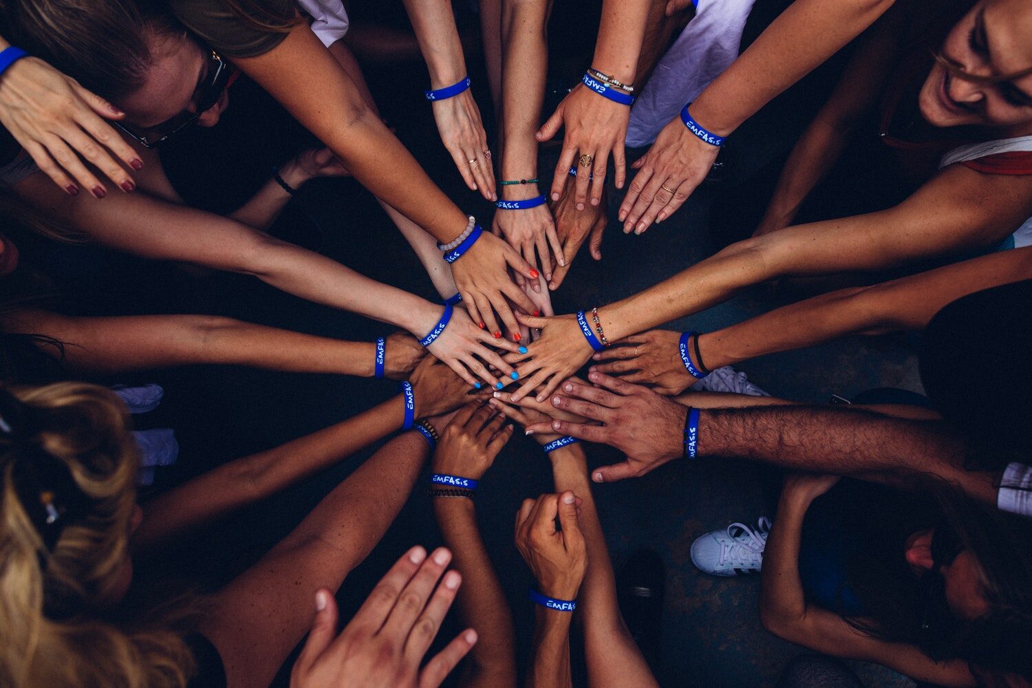 A circle of many people wearing “Emfasis” bracelets outstretch their hands to meet in the center of the circle.