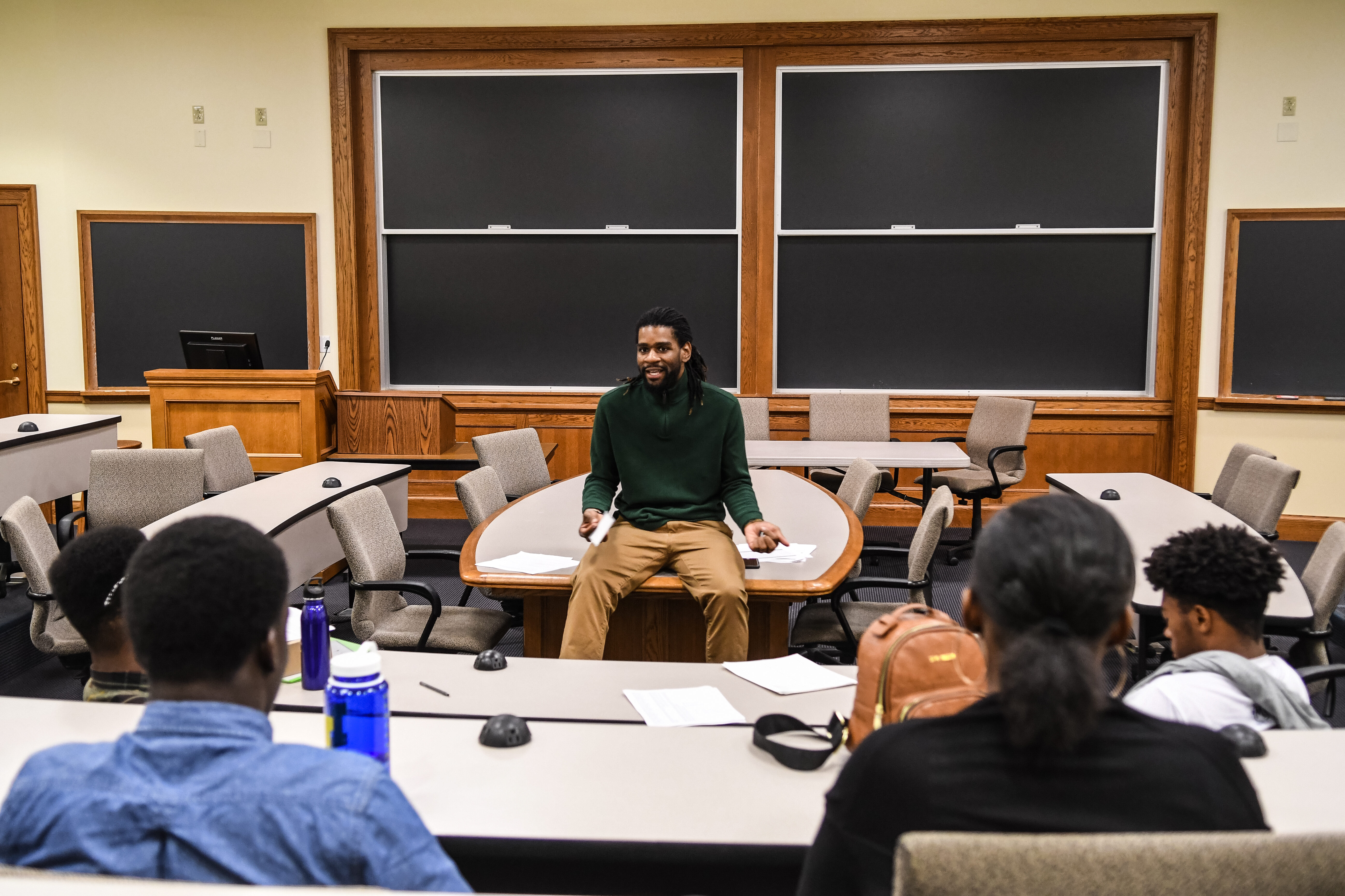 An instructor (shown from front) sits on a table, addressing students (shown from back) sitting at desks across from him.