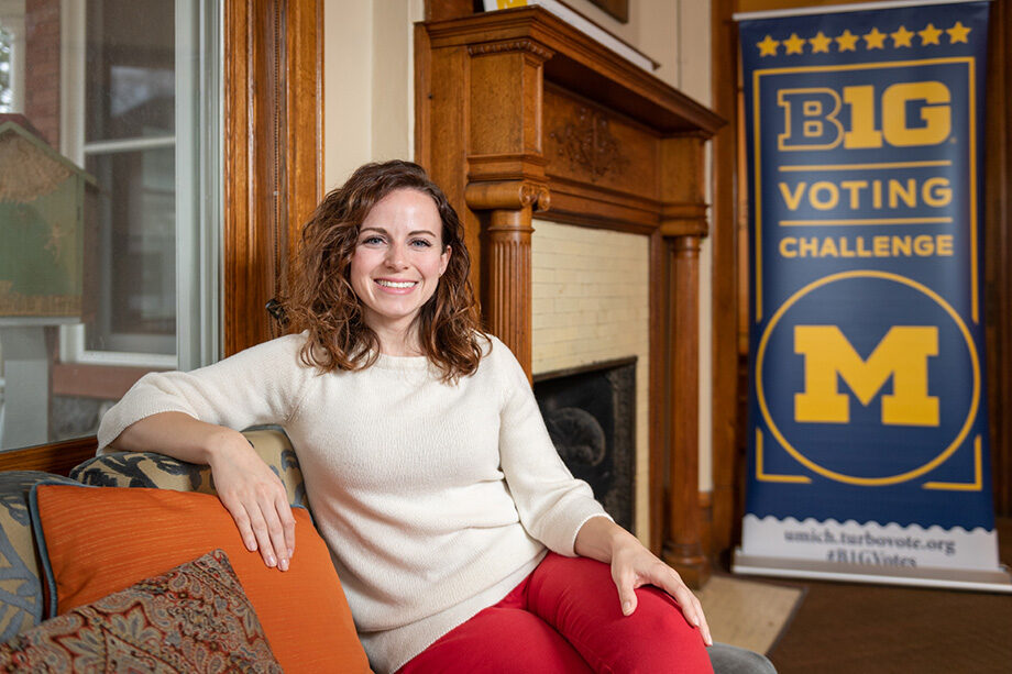 Erin Byrnes sits on a couch in the Ginsberg Center, smiling at the camera with a Big 10 Voting Challenge sign in the background.