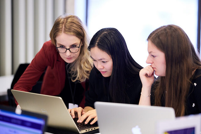 Three women sit and intently look at a laptop.