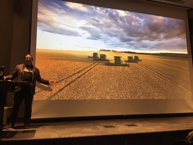 Sherman gestures to a screen showing a picture of machines plowing a field.