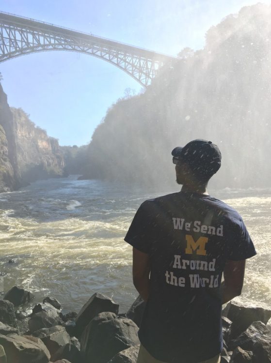 A young man sits in front of a mountainous landscape wearing a shirt that reads "We send M around the world."