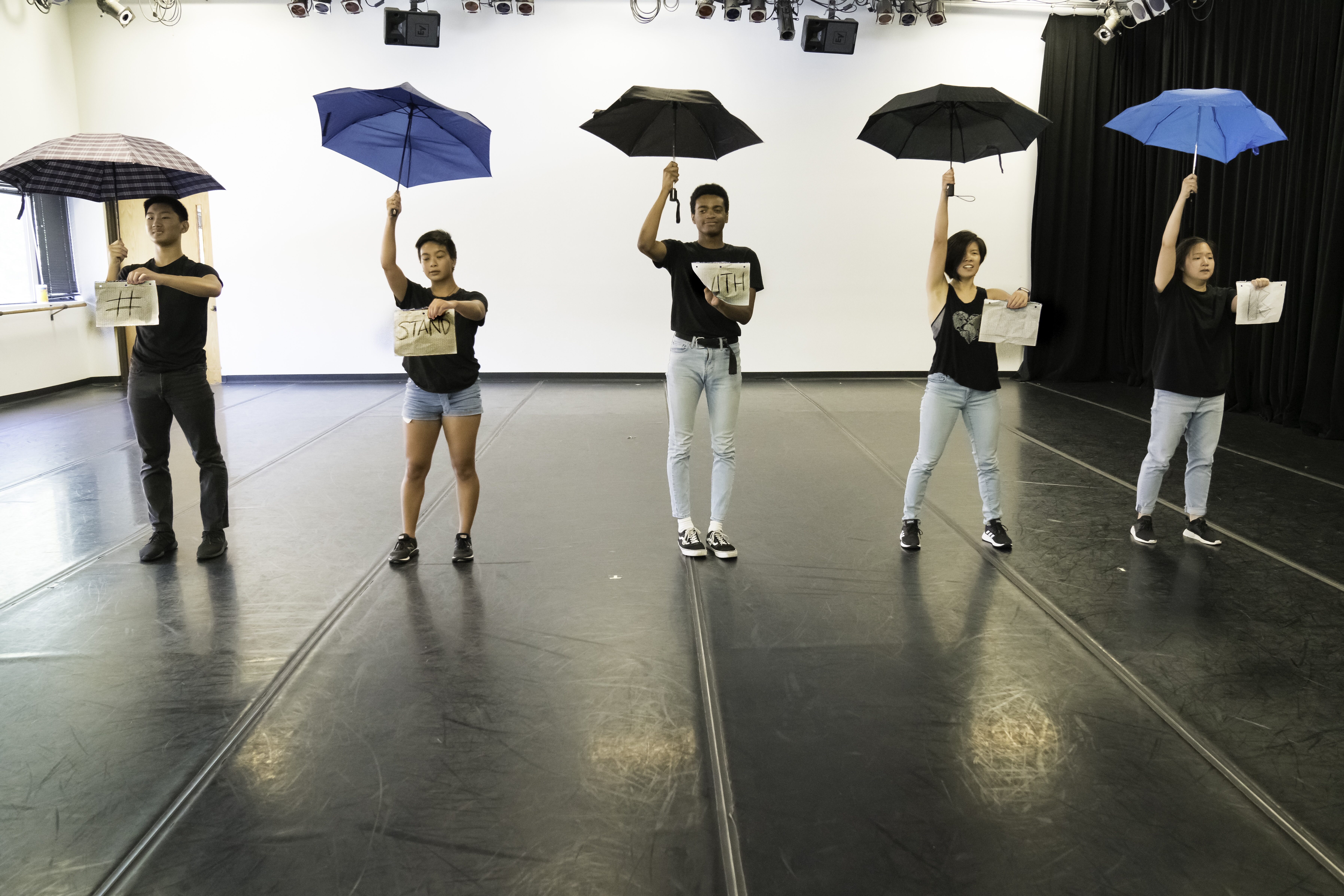 Four students stand with signs in front of them on one hand and umbrellas open and up in the other hand on a practice stage.